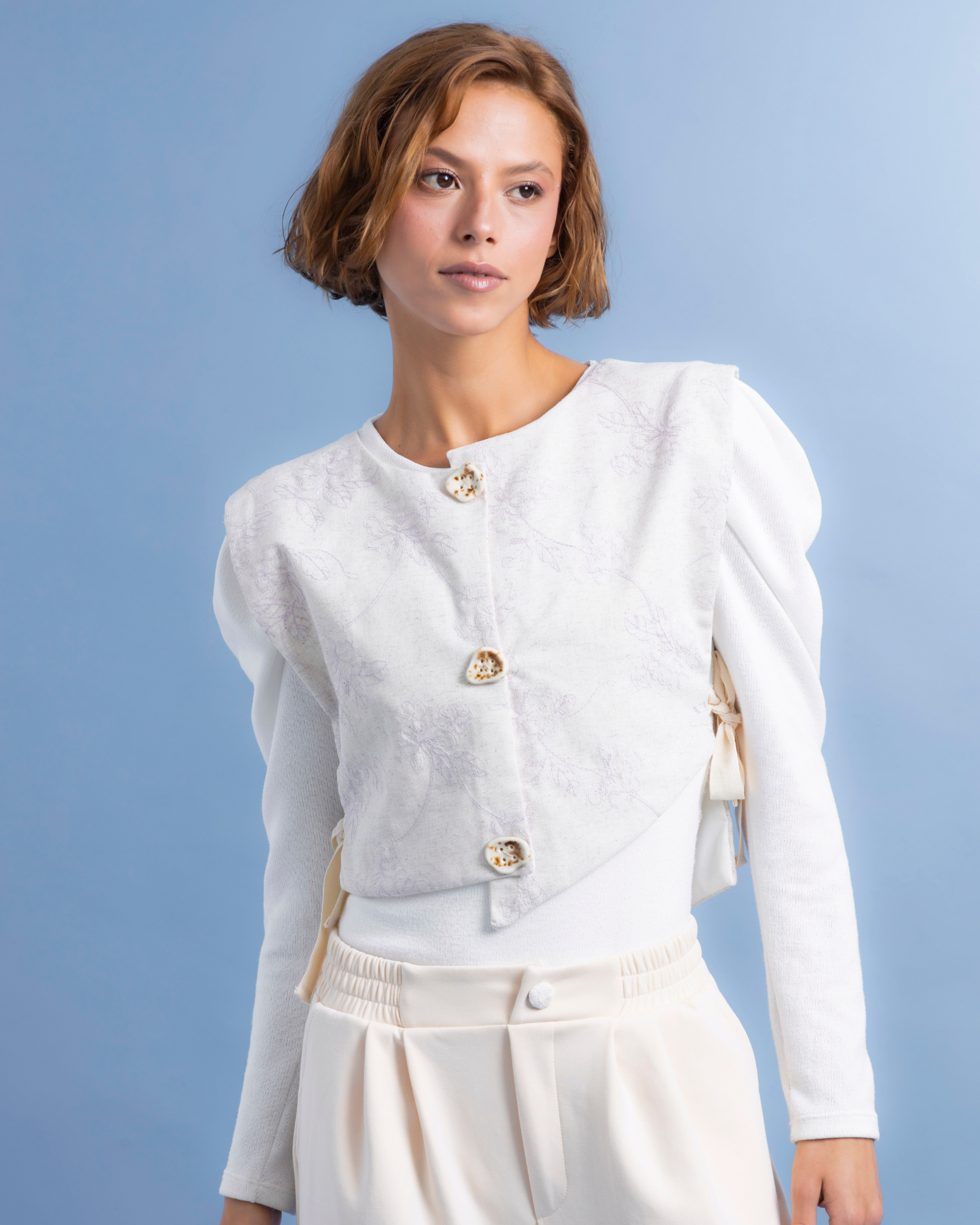 Chaleco tipo peto confeccionado en lino con textura de bordado, de estilo asimétrico. En el frente, botones decorativos de cerámica 100% hechos a mano.